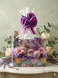 He Gives Strength Deluxe Wrapped Floral Spa Gift Basket in multicolor floral tray with Wonderful spa essentials and faith card wrapped in polka dot cellophane with purple magenta metallic pull bow surrounded by cream roses eucalyptus and lavender